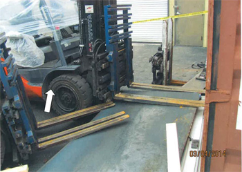 Photo of incident scene with forklift, forks lowered, with steel plate on ground in front of forklift, shipping container on far right of photo, in front of forklift.